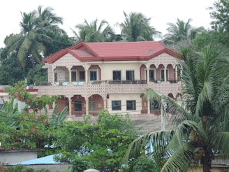 A two-story house with a red roof and intricate patterns on the exterior walls, surrounded by lush green palm trees and other foliage. The windows are large and dark, providing a modern contrast to the traditional design of the house.