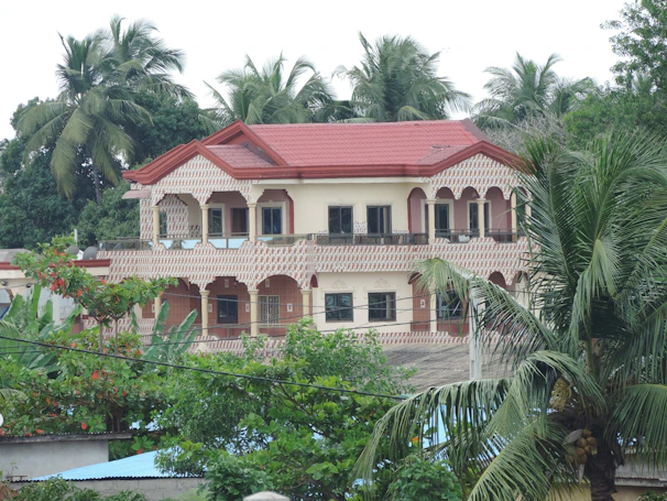 A two-story house with a red roof and intricate patterns on the exterior walls, surrounded by lush green palm trees and other foliage. The windows are large and dark, providing a modern contrast to the traditional design of the house.