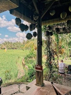 A rustic outdoor setting features a wooden structure with suspended decorations and a red sign indicating a toilet. The view includes lush green rice paddies under a partly cloudy sky. There are two people sitting at a table, surrounded by tropical vegetation.