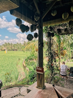 A rustic outdoor setting features a wooden structure with suspended decorations and a red sign indicating a toilet. The view includes lush green rice paddies under a partly cloudy sky. There are two people sitting at a table, surrounded by tropical vegetation.