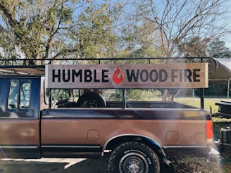 A truck with a wooden sign on its side that reads 'HUMBLE WOOD FIRE.' The truck is parked outdoors surrounded by trees with some branches devoid of leaves. Sunlight is filtering through the branches, casting shadows and creating a slightly warm and rustic atmosphere.