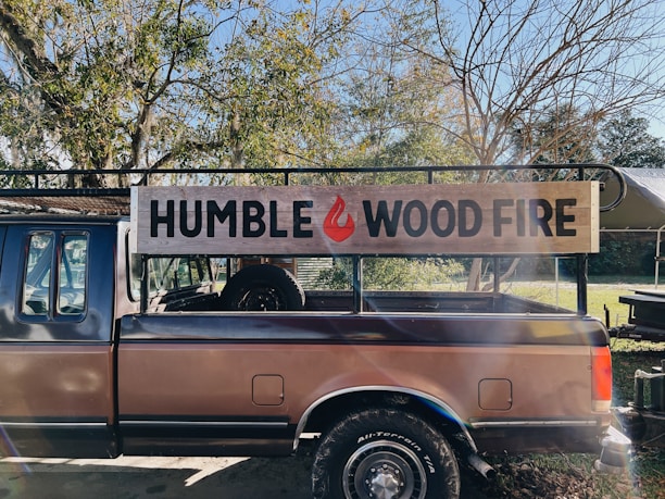 A truck with a wooden sign on its side that reads 'HUMBLE WOOD FIRE.' The truck is parked outdoors surrounded by trees with some branches devoid of leaves. Sunlight is filtering through the branches, casting shadows and creating a slightly warm and rustic atmosphere.
