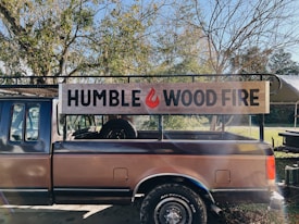 A truck with a wooden sign on its side that reads 'HUMBLE WOOD FIRE.' The truck is parked outdoors surrounded by trees with some branches devoid of leaves. Sunlight is filtering through the branches, casting shadows and creating a slightly warm and rustic atmosphere.