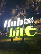 A brightly lit sign with the words 'Hub food town' and 'bite' in luminous letters stands against a dark background of tall palm trees and a building. The sign is illuminated with white and neon green colors, creating a vibrant contrast with the nighttime setting.
