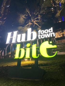 A brightly lit sign with the words 'Hub food town' and 'bite' in luminous letters stands against a dark background of tall palm trees and a building. The sign is illuminated with white and neon green colors, creating a vibrant contrast with the nighttime setting.