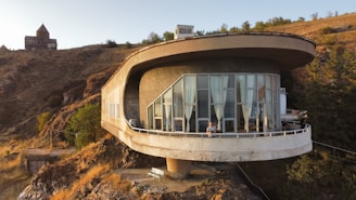 A modern, circular house with large windows is perched on a hillside. It has a futuristic design with a wide, overhanging roof and is surrounded by a rocky landscape with sparse vegetation. In the background, an old stone building, possibly a church, is visible atop another hill. The scene is set during sunset, casting a warm golden glow.