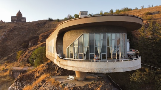 A modern, circular house with large windows is perched on a hillside. It has a futuristic design with a wide, overhanging roof and is surrounded by a rocky landscape with sparse vegetation. In the background, an old stone building, possibly a church, is visible atop another hill. The scene is set during sunset, casting a warm golden glow.