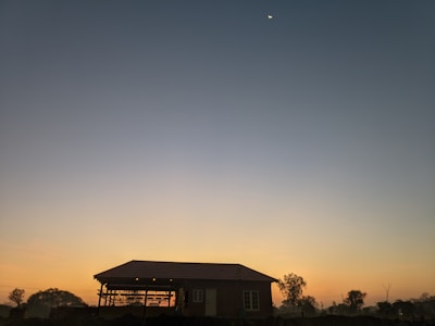 A small house with a sloped roof is silhouetted against a twilight sky. The sky transitions from a warm orange near the horizon to a deeper blue as it rises. A bright crescent moon is visible in the upper part of the sky, and there are several trees around the house.