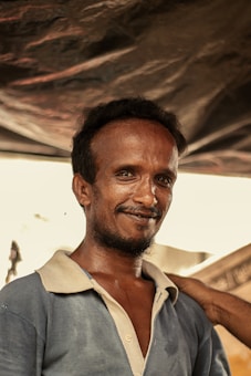 A man with a short beard and mustache is smiling slightly. He is wearing a blue collared shirt with a beige inner lining. His hair is short and curly. There is a hand resting on his shoulder in a supportive gesture. The background appears to be under some kind of canopy or tent, with light coming through in some areas creating a soft, natural lighting effect.