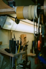 A workbench setting featuring various tools neatly organized on a wooden shelf. The tools include a diverse array of handles and blades, some sharp and pointed, with a collection of tape rolls stored above them. Overhead lighting illuminates the area, casting soft shadows.