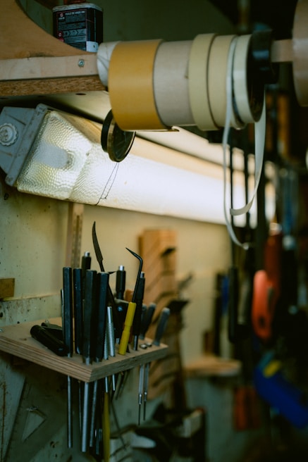 A workbench setting featuring various tools neatly organized on a wooden shelf. The tools include a diverse array of handles and blades, some sharp and pointed, with a collection of tape rolls stored above them. Overhead lighting illuminates the area, casting soft shadows.