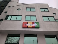 The image depicts a modern building with large green-tinted windows. A colorful sign with the word 'mBank' is prominently mounted near the center of the facade. The building's architecture features geometric shapes and a light gray exterior.
