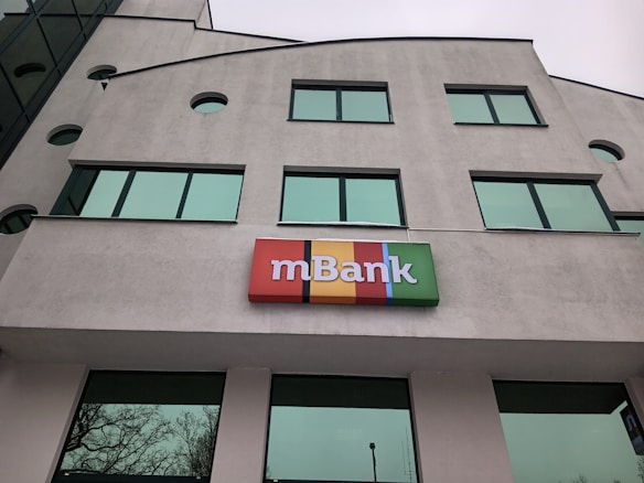 The image depicts a modern building with large green-tinted windows. A colorful sign with the word 'mBank' is prominently mounted near the center of the facade. The building's architecture features geometric shapes and a light gray exterior.