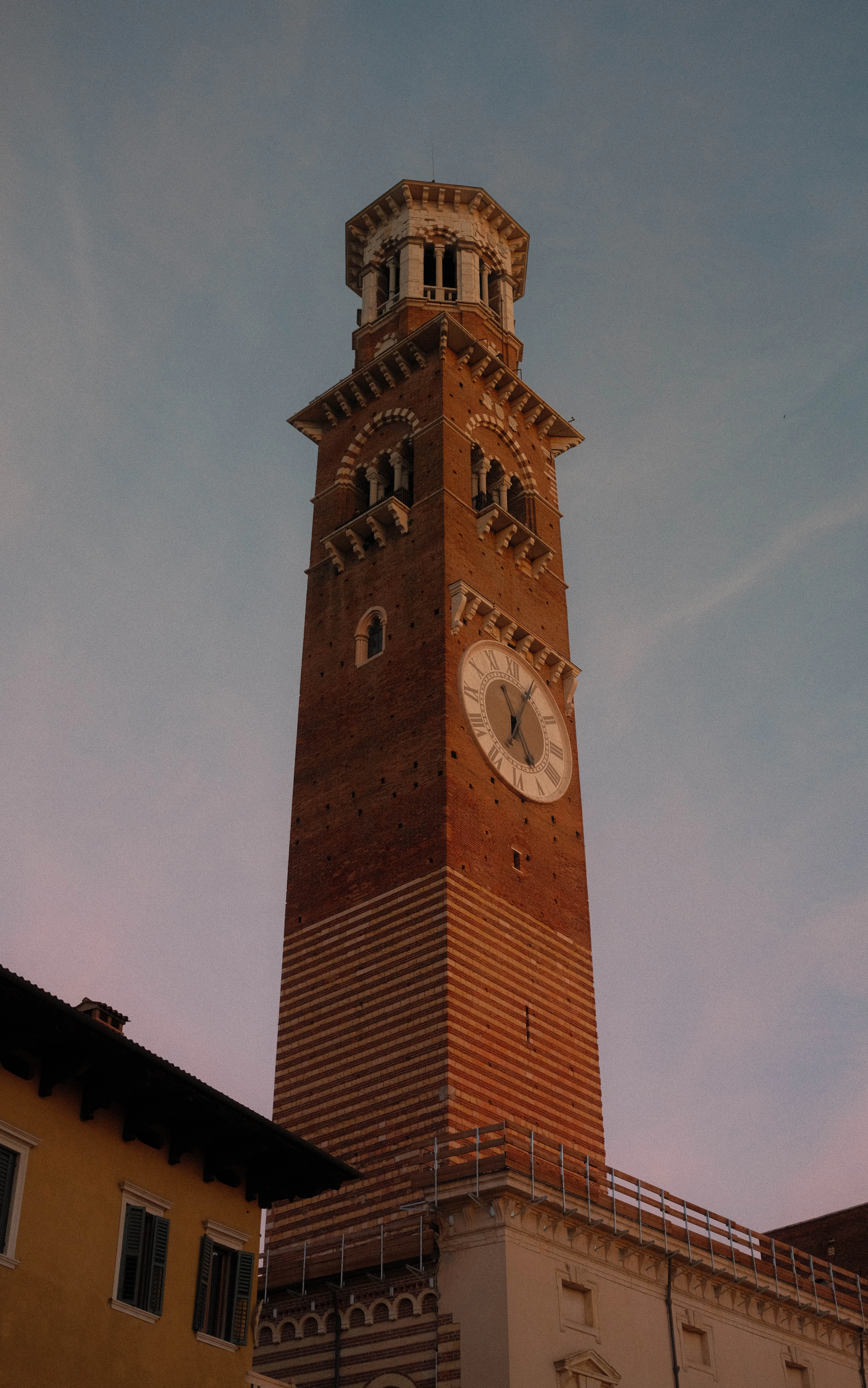 The banded brick and tufa shaft of Verona's Torre dei Lamberti rising to its white marble belfry and round clock face against a pale blue and pink dusk sky.