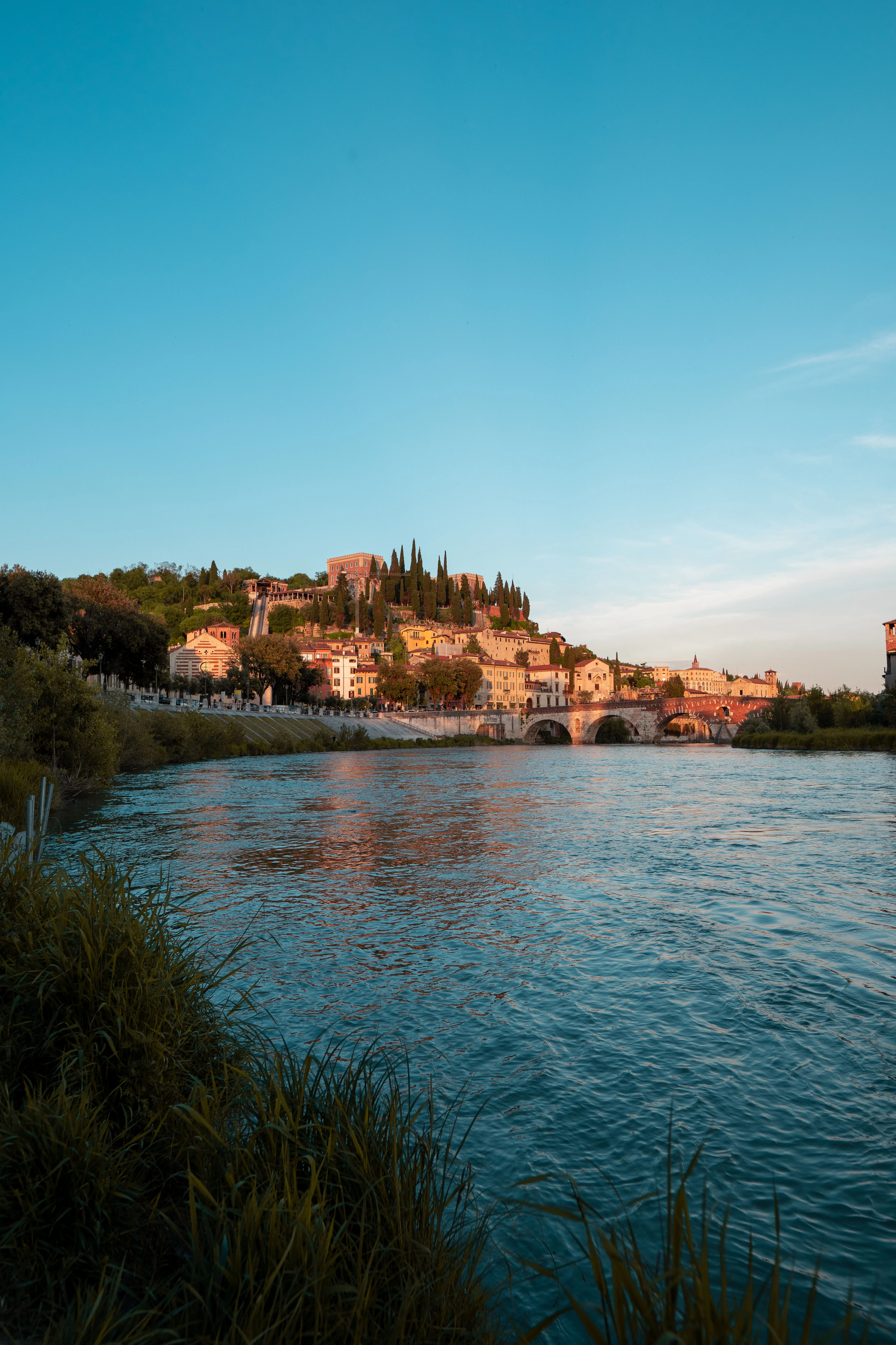 Castel San Pietro's cypress-covered hillside glows at sunset above the Adige River and a stone bridge.