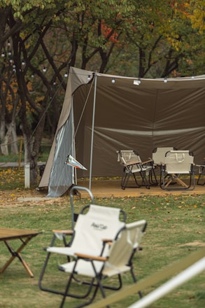 A camping scene features a large, open canopy tent with foldable chairs arranged on a wooden platform underneath. The area is surrounded by trees with leaves showing autumn colors. String lights hang from the tent, adding a cozy atmosphere. A grassy area with scattered leaves and small tables is visible in the foreground.
