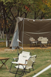 A camping scene features a large, open canopy tent with foldable chairs arranged on a wooden platform underneath. The area is surrounded by trees with leaves showing autumn colors. String lights hang from the tent, adding a cozy atmosphere. A grassy area with scattered leaves and small tables is visible in the foreground.