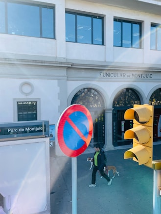 A man walking a dog near the entrance of the Funicular de Montjuïc. A traffic light and no parking sign are visible, with Parc de Montjuïc signage nearby. The building in the background has multiple windows and architectural features like arches.