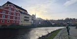 A scenic view of a European town by a river during sunset. The image features colorful buildings, including a prominent red building with the words 'hotel mlýn' painted on it. A person is seen walking along the cobblestone path by the riverbank, carrying a green backpack. The sky has a soft gradient from light to dark with subtle cloud patterns.