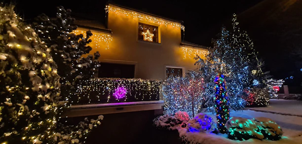 A cozy house glowing with colorful Christmas lights wrapped around trees and lining the pathway.