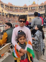 Two children are standing among a crowd in an outdoor stadium or arena. The children are holding Indian flags and have the Indian tricolor painted on their faces. The background shows a large structure with arches, filled with many spectators, and there is a banner indicating it is a venue in India.