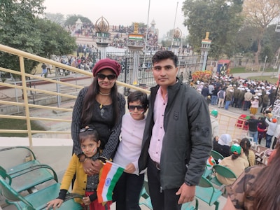 A family with two children stands in front of a crowded area. The adults and children have painted faces and are holding an Indian flag. In the background, there are many people gathered, some wearing uniforms or hats, and a decorative gate is visible with flags and symbols.