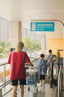 People are seen using a moving walkway while pushing shopping trolleys at an IKEA store. The walkway has a sign above it indicating that it is only for IKEA shopping trolleys, with a weight limit of 130kg. The setting is modern and urban, with a view of buildings and trees in the background.