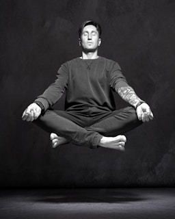 A person is in a meditative pose, appearing to levitate. The individual sits cross-legged with eyes closed, exuding a sense of tranquility. The right arm is visible, with a tattoo, and the hands are open with palms facing upwards. The image is in black and white, creating a dramatic contrast.