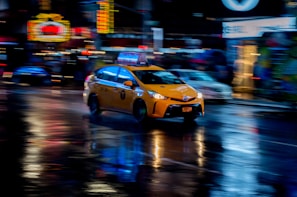 A yellow taxi is moving quickly through a city street at night. Neon lights and signs are blurred in the background, reflecting off the wet pavement, creating a dynamic and vibrant atmosphere.