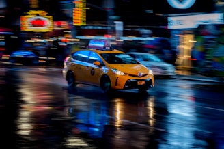 A yellow taxi is moving quickly through a city street at night. Neon lights and signs are blurred in the background, reflecting off the wet pavement, creating a dynamic and vibrant atmosphere.