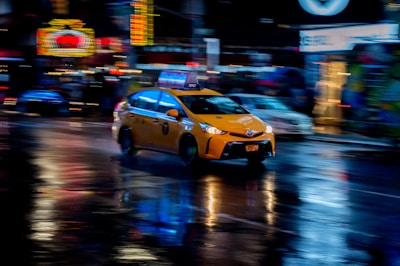 A yellow taxi is moving quickly through a city street at night. Neon lights and signs are blurred in the background, reflecting off the wet pavement, creating a dynamic and vibrant atmosphere.