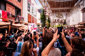A lively crowd gathers in an outdoor area with people holding up smartphones, apparently capturing the moment. Festive decorations hang above, and buildings with brick and stucco facades can be seen. Some individuals are waving flags, creating a celebratory atmosphere.