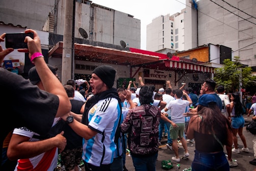 A crowd of enthusiastic people wearing sports jerseys and casual clothing are gathered outside a building with a red and white striped awning. Many have their arms raised in celebration, and there appears to be some sort of festive activity happening with liquids being sprayed into the air.