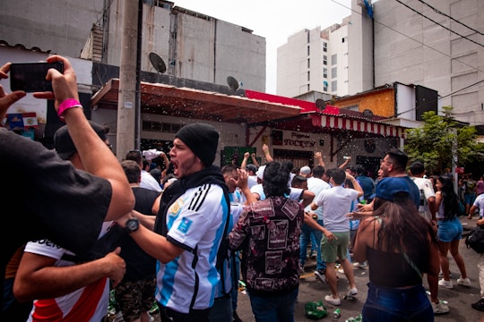 A crowd of enthusiastic people wearing sports jerseys and casual clothing are gathered outside a building with a red and white striped awning. Many have their arms raised in celebration, and there appears to be some sort of festive activity happening with liquids being sprayed into the air.