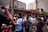 A crowd of enthusiastic people wearing sports jerseys and casual clothing are gathered outside a building with a red and white striped awning. Many have their arms raised in celebration, and there appears to be some sort of festive activity happening with liquids being sprayed into the air.