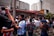 A crowd of enthusiastic people wearing sports jerseys and casual clothing are gathered outside a building with a red and white striped awning. Many have their arms raised in celebration, and there appears to be some sort of festive activity happening with liquids being sprayed into the air.