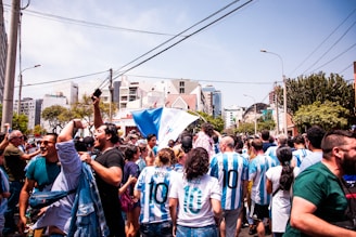 A large group of people fills the street in a celebratory atmosphere, with many wearing sports jerseys featuring the number 10 and the name 'Messi'. Flags are waved enthusiastically in the air. The background displays urban elements such as buildings, trees, and street lights under a clear blue sky.