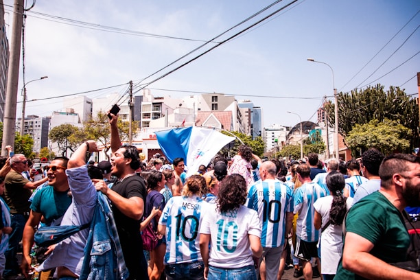 A large group of people fills the street in a celebratory atmosphere, with many wearing sports jerseys featuring the number 10 and the name 'Messi'. Flags are waved enthusiastically in the air. The background displays urban elements such as buildings, trees, and street lights under a clear blue sky.