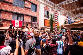 A lively crowd of people gathered together outdoors, many holding up smartphones to capture an event. A red and white flag with a crest hangs from a brick building in the background. The atmosphere seems festive, with people dressed casually, some wearing hats and jerseys. There are colorful umbrellas and decorative elements above suggesting a celebration or gathering.