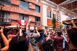 A lively outdoor gathering features a crowd of people raising their hands and phones in celebration. The backdrop includes a red brick building adorned with flowering plants and a flag. Light streams in, suggesting a festive atmosphere in a sunny courtyard.