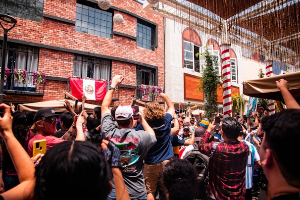 A lively outdoor gathering features a crowd of people raising their hands and phones in celebration. The backdrop includes a red brick building adorned with flowering plants and a flag. Light streams in, suggesting a festive atmosphere in a sunny courtyard.