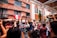 A lively outdoor gathering features a crowd of people raising their hands and phones in celebration. The backdrop includes a red brick building adorned with flowering plants and a flag. Light streams in, suggesting a festive atmosphere in a sunny courtyard.