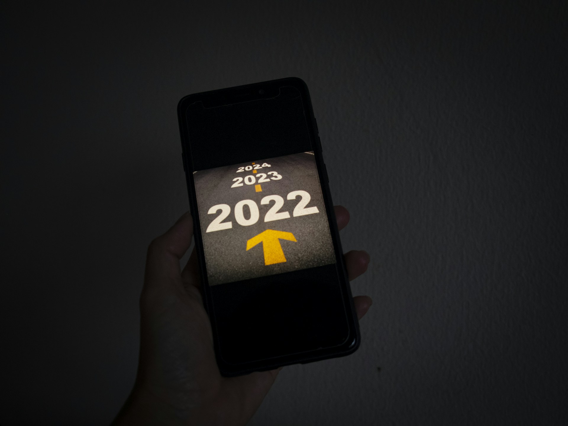 A hand holds a smartphone displaying the years 2022, 2023, and 2024 on a dark background, with a yellow arrow pointing forward from 2022 toward 2023 and 2024.
