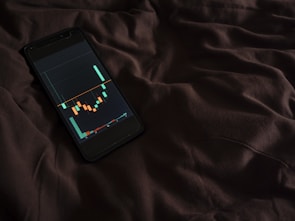 A smartphone displaying a financial chart with candlestick patterns is placed on a dark, crumpled fabric surface. The chart features various colored candlesticks against a dark background, suggesting financial analysis or stock market activity.