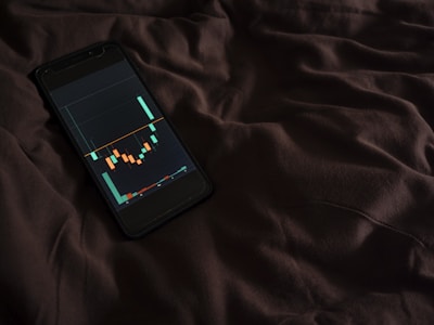 A smartphone displaying a financial chart with candlestick patterns is placed on a dark, crumpled fabric surface. The chart features various colored candlesticks against a dark background, suggesting financial analysis or stock market activity.