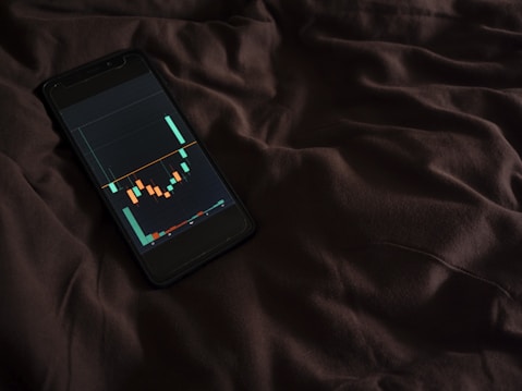 A smartphone displaying a financial chart with candlestick patterns is placed on a dark, crumpled fabric surface. The chart features various colored candlesticks against a dark background, suggesting financial analysis or stock market activity.