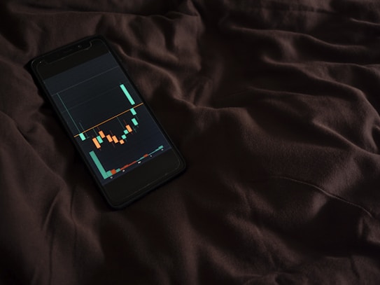 A smartphone displaying a financial chart with candlestick patterns is placed on a dark, crumpled fabric surface. The chart features various colored candlesticks against a dark background, suggesting financial analysis or stock market activity.