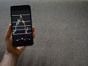 A hand is holding a smartphone that displays a stock market chart with various candlestick patterns and horizontal lines. The phone is held against a textured fabric background, which appears to be a sofa or carpet.