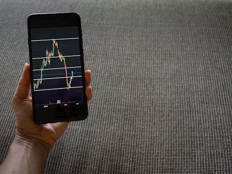 A hand is holding a smartphone that displays a stock market chart with various candlestick patterns and horizontal lines. The phone is held against a textured fabric background, which appears to be a sofa or carpet.