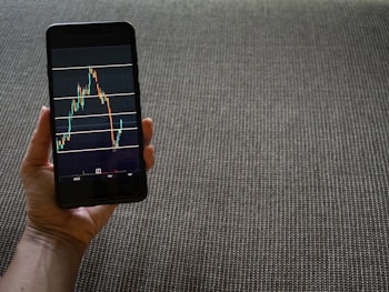 A hand is holding a smartphone that displays a stock market chart with various candlestick patterns and horizontal lines. The phone is held against a textured fabric background, which appears to be a sofa or carpet.